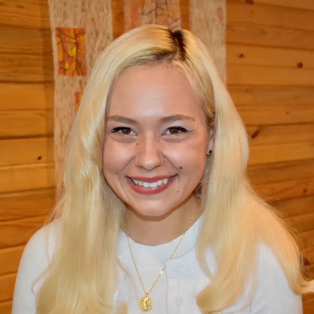 Arionna Timpson smiling in a white blouse, against a warm wood-slat wall.